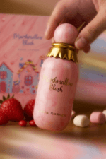 Main tenant le flacon rose marbré et doré du parfum Marshmallow Blush de Paris Corner avec fraises et marshmallows en arrière-plan