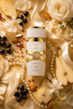 Vue de dessus du flacon Lattafa Yara Moi blanc et or couché parmi des roses blanches, jasmin, tranches de poire, cassis, poivre rose, amandes, collier de perles et ruban satiné ivoire sur fond doré